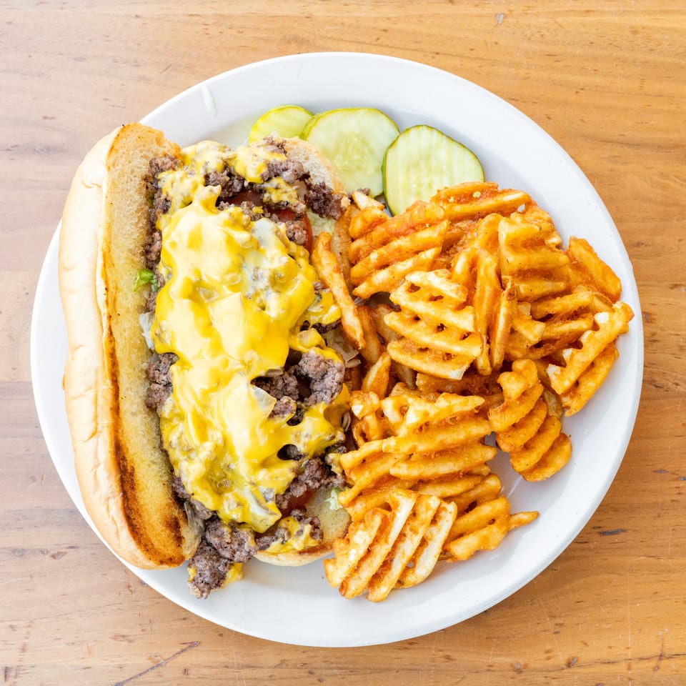 Cheesesteak sandwich with melted yellow cheese, waffle fries, and pickle slices on a white plate.