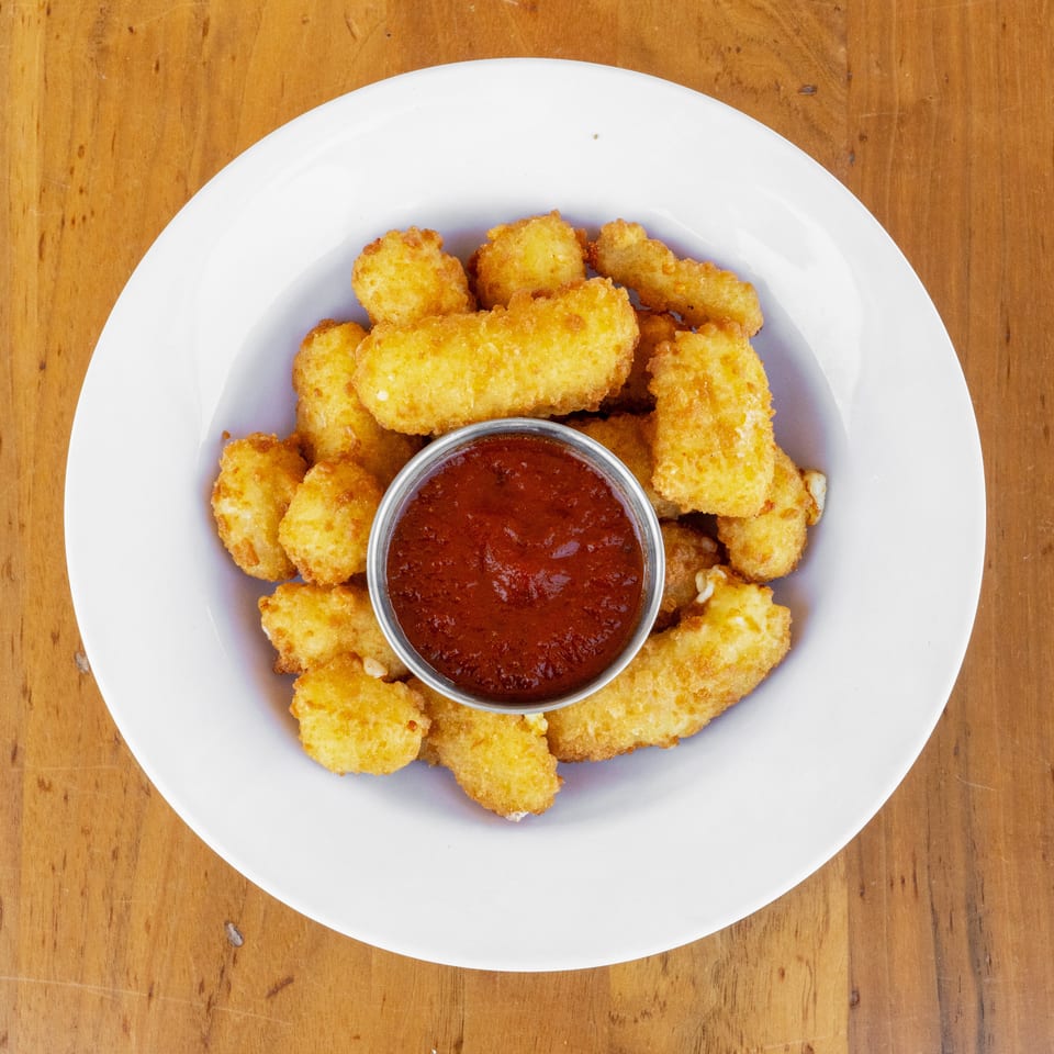 Golden fried cheese bites arranged around a cup of red dipping sauce on a white plate atop a wooden table.